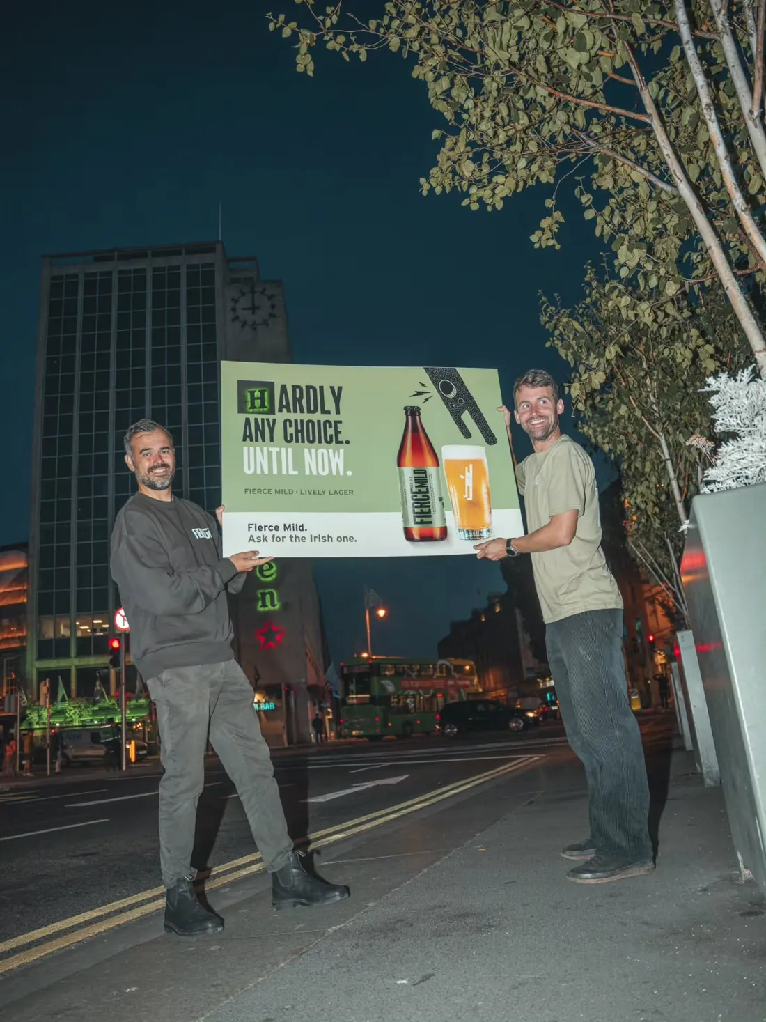 Fergal and Cathal holding a Fierce Mild billboard on O'Connell Bridge at night, lined up so the Heineken sign sits behind it
